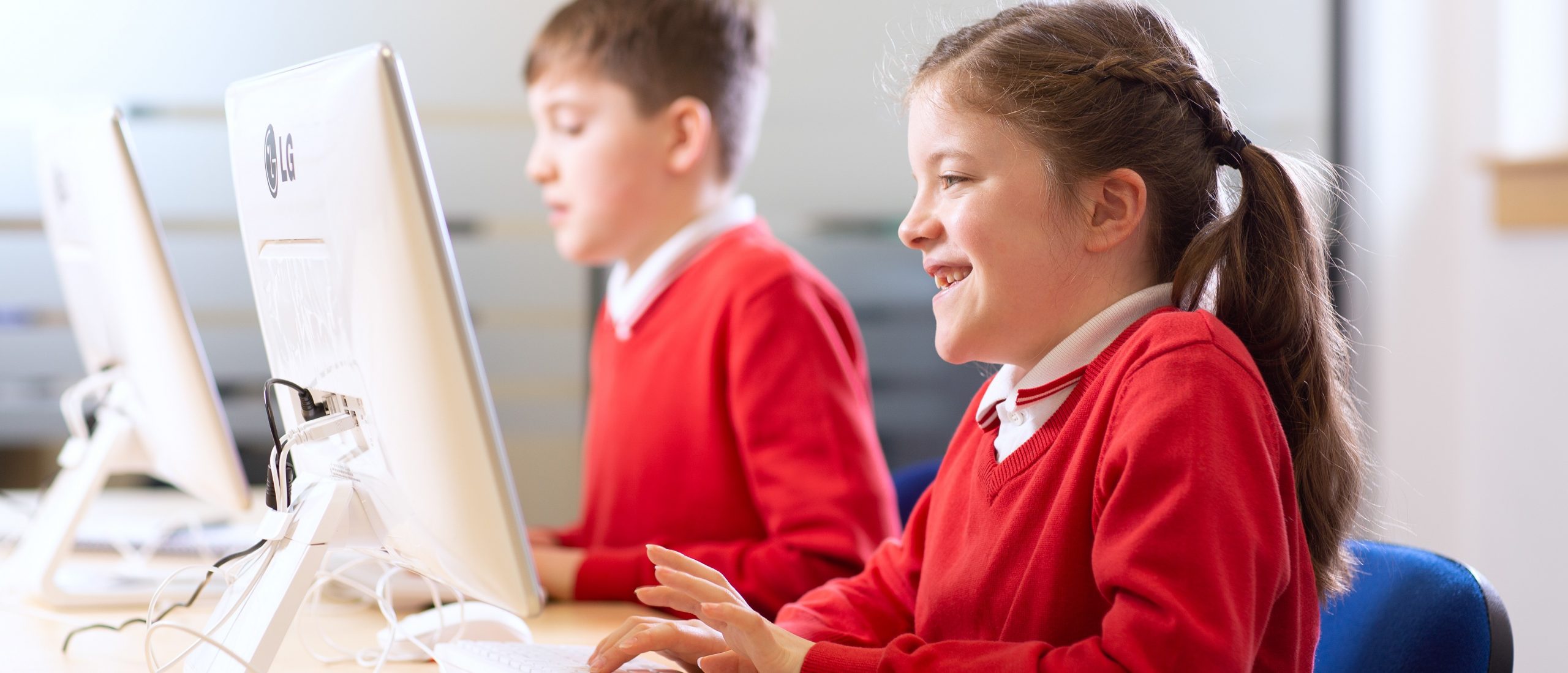 Primary school-age girl sat at a desktop computer, smiling while using Lexia literacy software for schools. A boy of the same age is sat at a computer in the background also using the software.